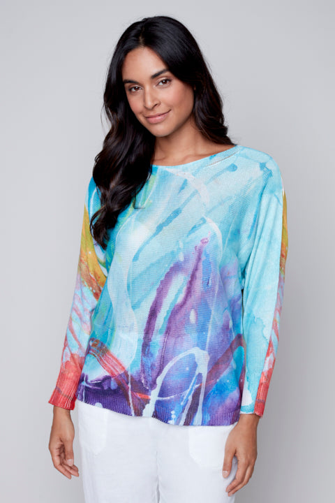 Woman in a colorful Claire Desjardins long sleeves abstract print top and white pants posing against a neutral background.