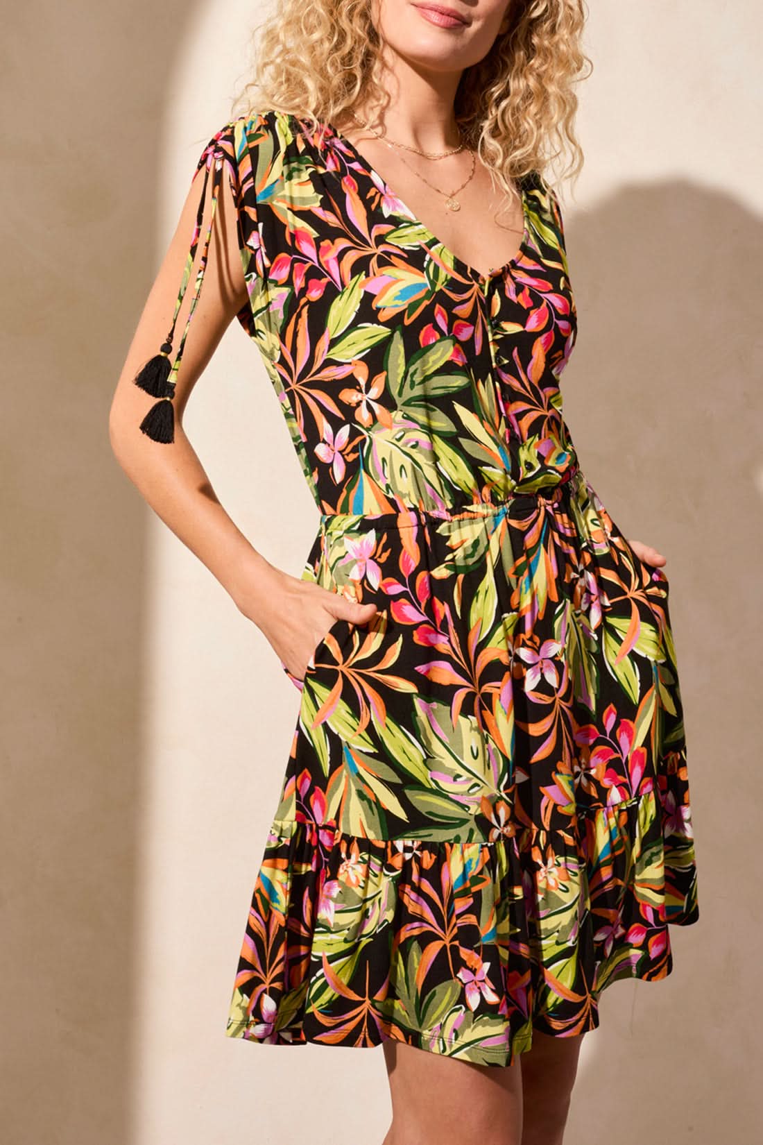 A woman wears the Tribal ADJUSTABLE CAP SLEEVES V NECK DRESS, a short, colorful floral dress with ruffled hem and adjustable cap sleeves, paired with brown high-heeled sandals against a light background.