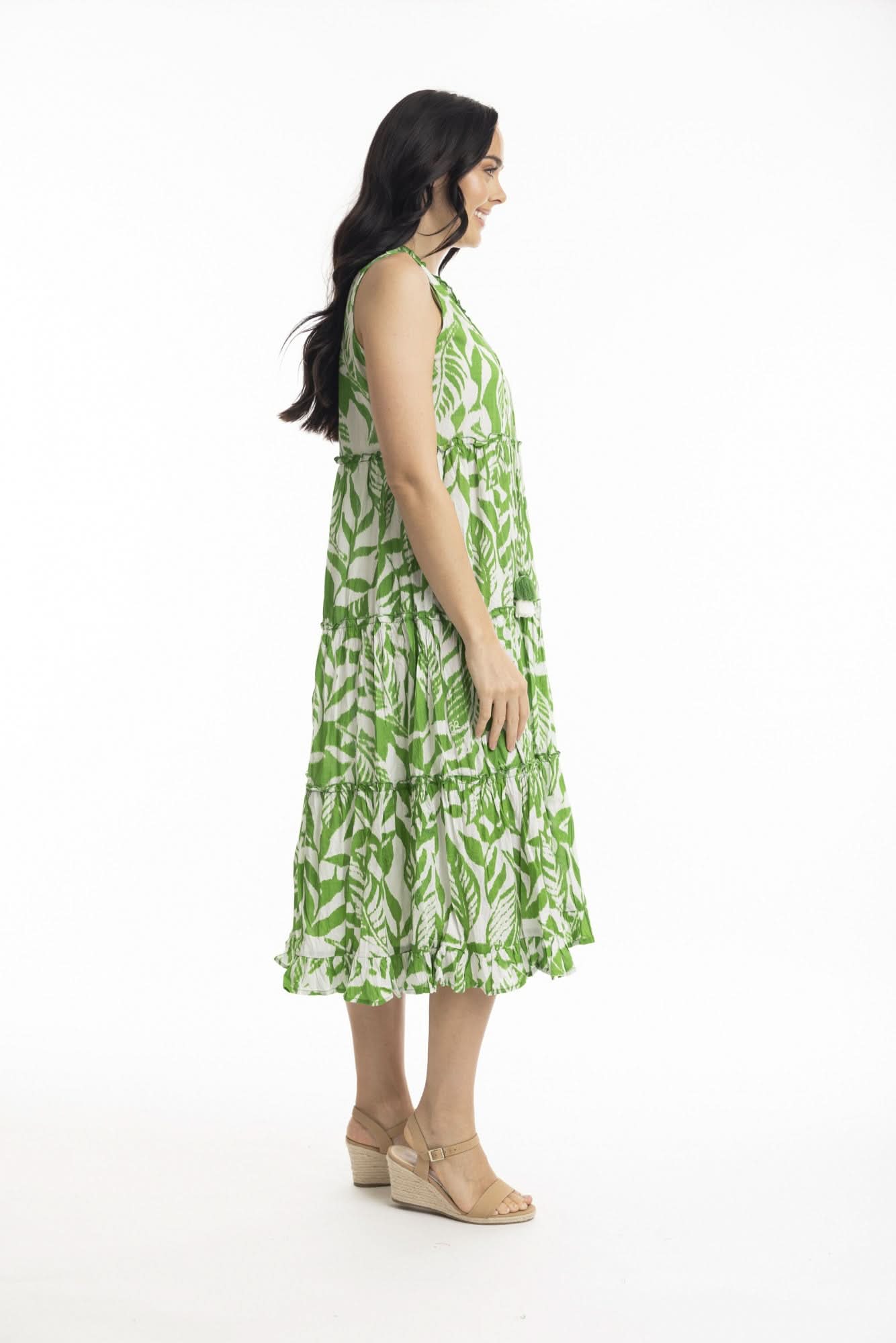 A woman smiles in the Orientique Hayman Green Sleeveless Boho Dress, featuring a green and white leaf pattern and tiered skirt, styled with beige heeled sandals—ideal for summer days against a plain white background.
