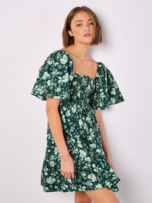 A person wearing the Apricot's Watercolor Rose Smocked Mini, a green floral dress with puffed sleeves, stands against a plain white background.