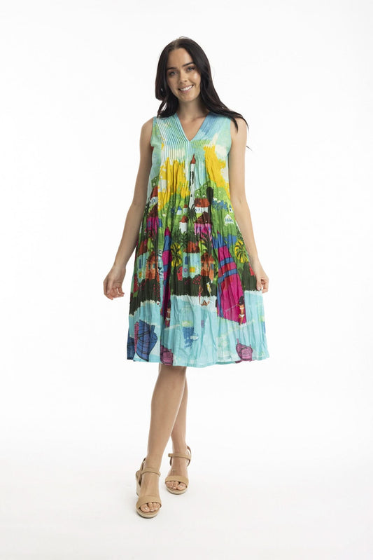 A woman stands in profile against a white background, wearing the Orientique Florida Cotton Pleated Dress with a vibrant cityscape pattern and V-neck—styled with nude wedge sandals for an effortlessly chic summer look.