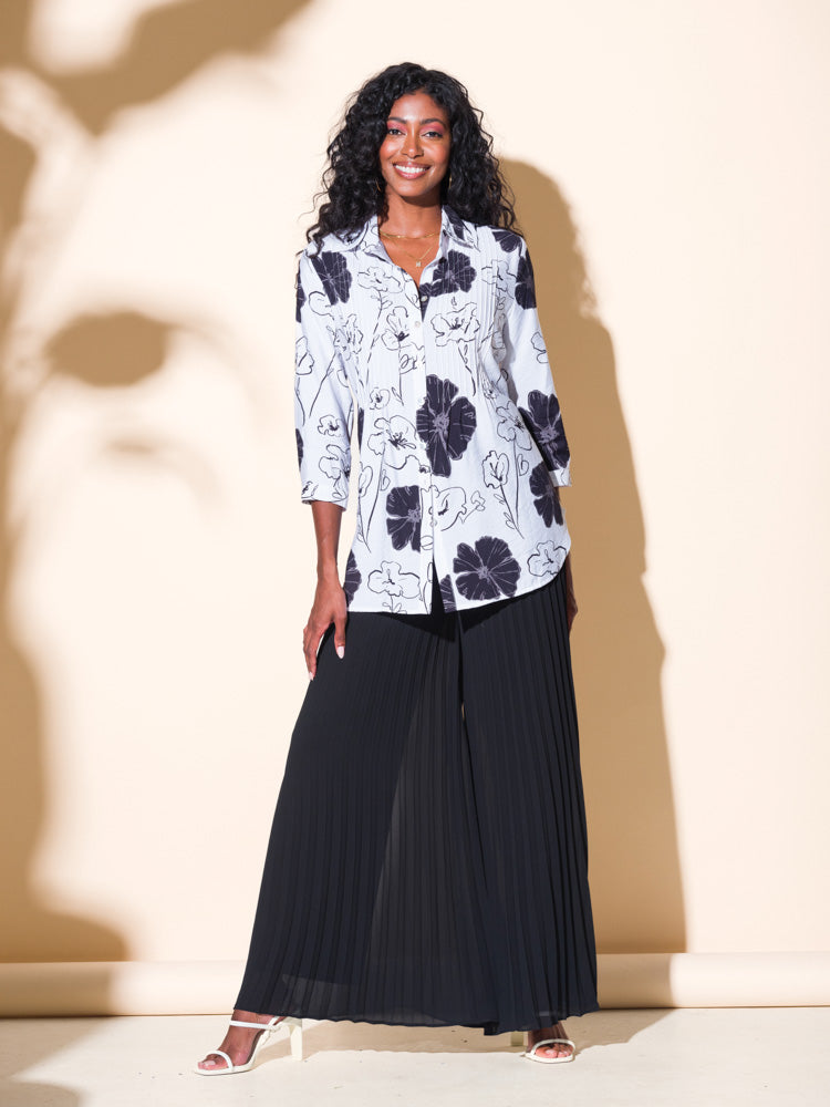 An elegant woman wearing black pleated pants and an Alison Sheri Black and White Button Up Floral Blouse showcases her impeccable style.
