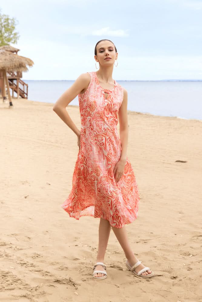 A woman stands on a sandy beach with huts and water in the background, wearing the Alison Sheri Paisley Sleeveless Dress featuring an elegant belted silhouette in orange and white.