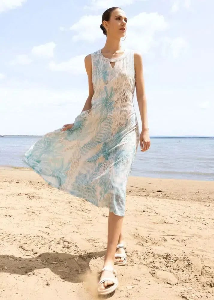 A woman in an Alison Sheri Flare Dress walks barefoot on a sandy beach near the water on a sunny day.