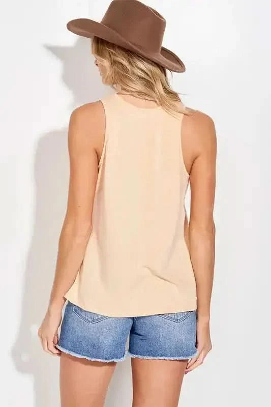 A woman in a "Aztec Print Country Trendy" tank top from Don't Be Chy Boutique, blue jeans, and a brown hat poses with her arms raised, touching the brim of the hat.