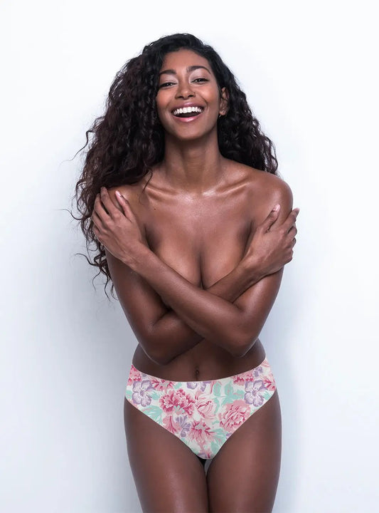 A smiling woman with curly hair covering her chest with her arms, wearing comfortable floral Body Hush 365 Bikini bottoms against a white background.