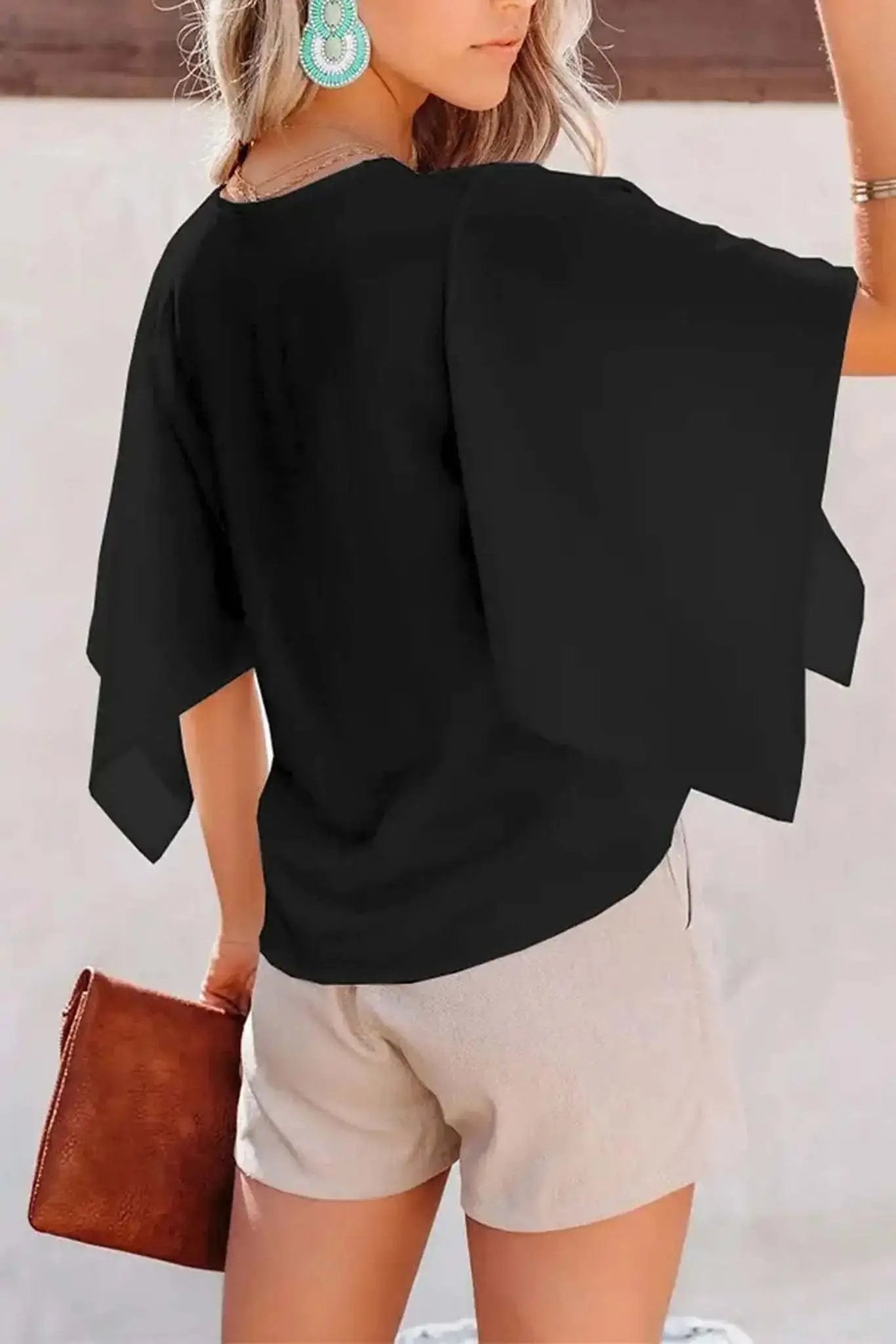 Woman in a black V-neck top with flare sleeves and distressed denim shorts holding a brown clutch from Don't Be Chy Boutique.