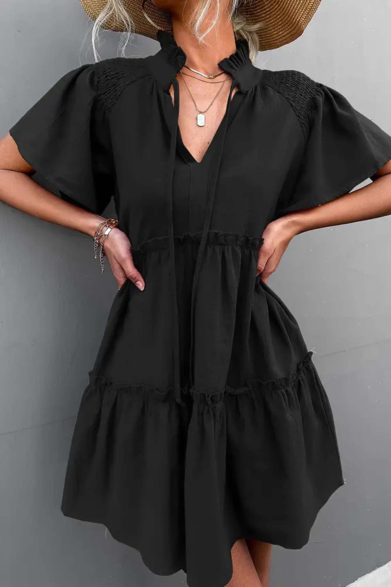 A woman in a black ruffled mini dress with puff sleeves and a tied neckline, holding the skirt, standing outdoors by Don't Be Chy Boutique.
