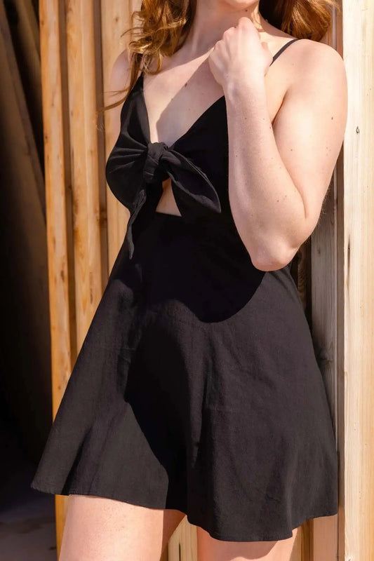 A woman wearing a Don't Be Chy Boutique Bow Knot Backless Sling Dress in black stands by a wooden fence, partially cropped to show from the neck down.