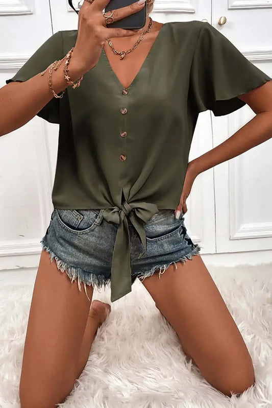 Woman wearing a green Don't Be Chy Boutique Button Down V Neck Front Tie Short Sleeves Top and denim shorts, accessorized with bracelets, taking a selfie. The visible background is white and fluffy.