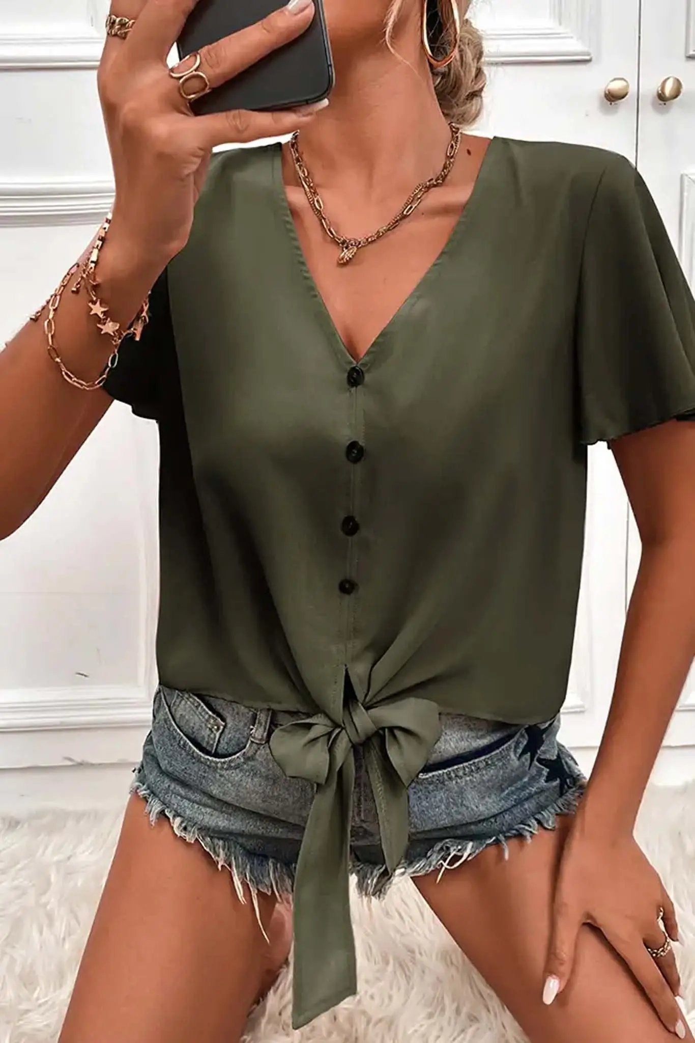 Woman wearing a green Don't Be Chy Boutique Button Down V Neck Front Tie Short Sleeves Top and denim shorts, accessorized with bracelets, taking a selfie. The visible background is white and fluffy.