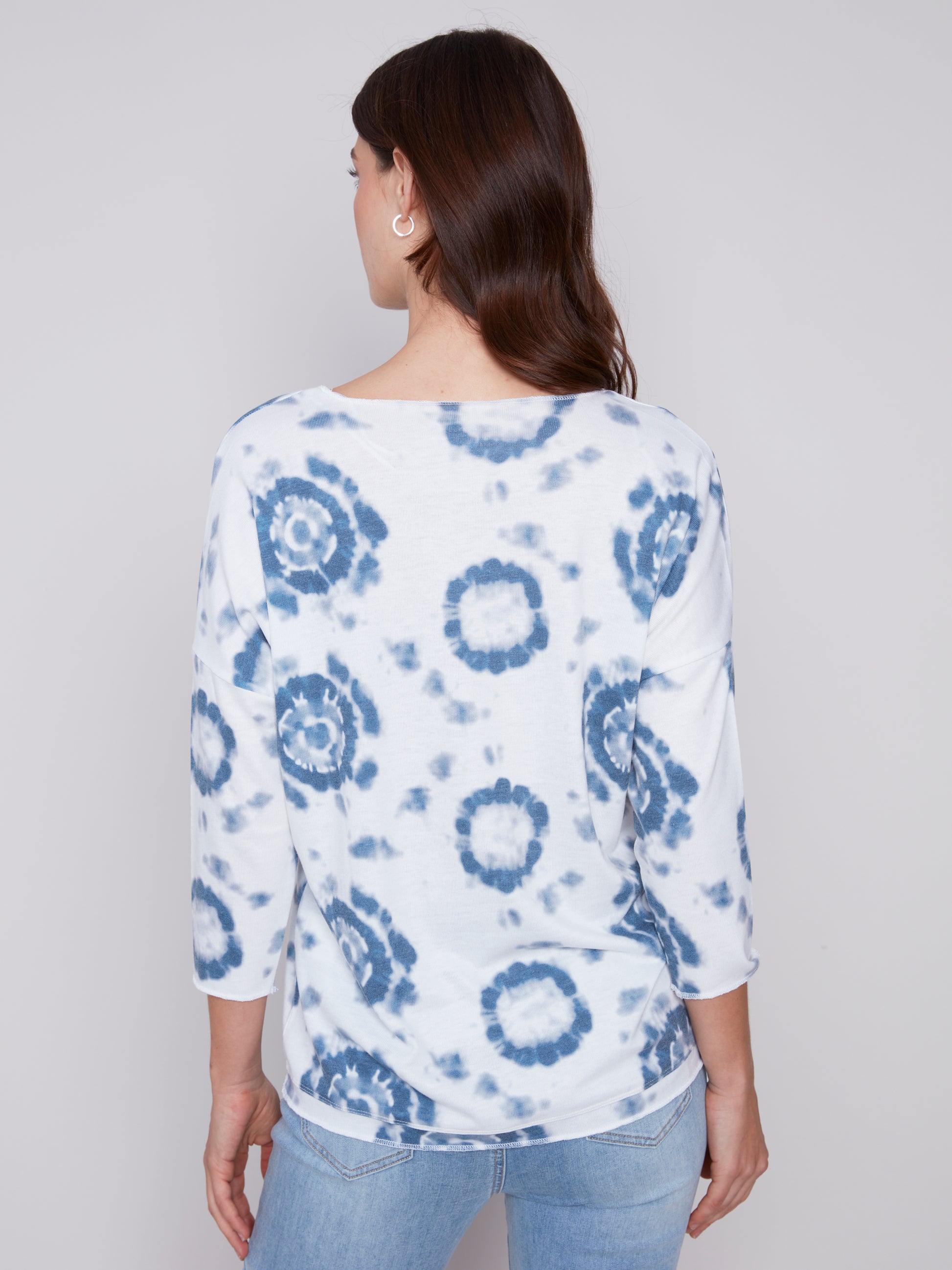 A woman in a stylish, Charlie B Indigo Printed V Neck Top and blue jeans standing against a neutral background.