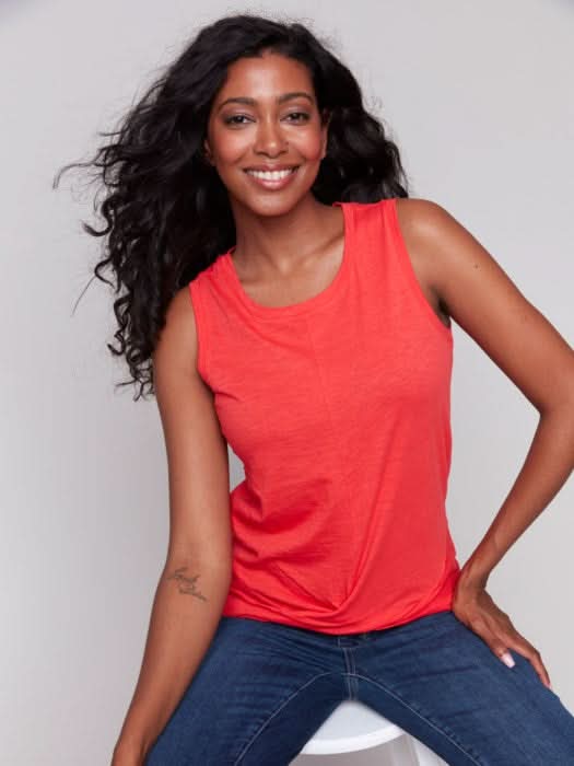 A woman with long, wavy dark hair wears the Charlie B Cotton Slub Tank Top With Knot Detail in red and blue jeans, standing against a light gray background while touching her hair.