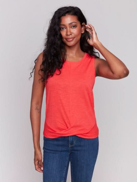 A woman with long, wavy dark hair wears the Charlie B Cotton Slub Tank Top With Knot Detail in red and blue jeans, standing against a light gray background while touching her hair.