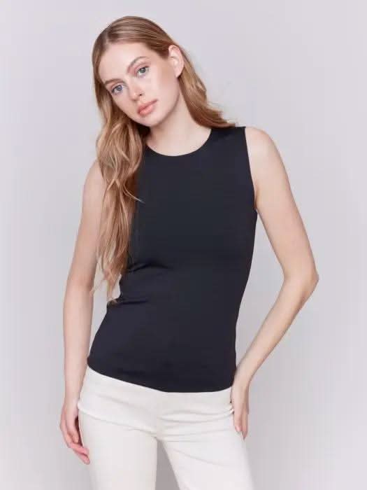 A person with long curly hair, wearing a Charlie B Super Stretch Sleeveless Top, blue jeans, and black ankle boots, stands posing against a plain background.