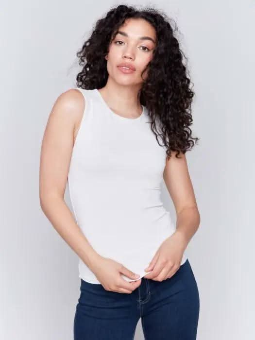 A person with long curly hair, wearing a Charlie B Super Stretch Sleeveless Top, blue jeans, and black ankle boots, stands posing against a plain background.