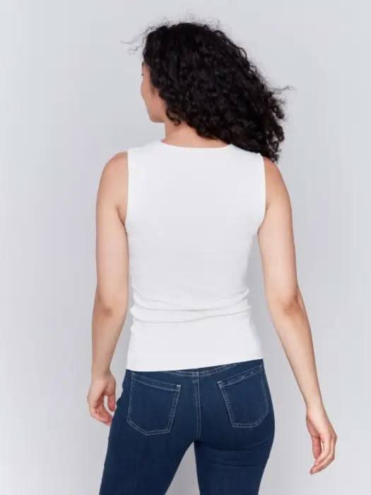 A person with long curly hair, wearing a Charlie B Super Stretch Sleeveless Top, blue jeans, and black ankle boots, stands posing against a plain background.