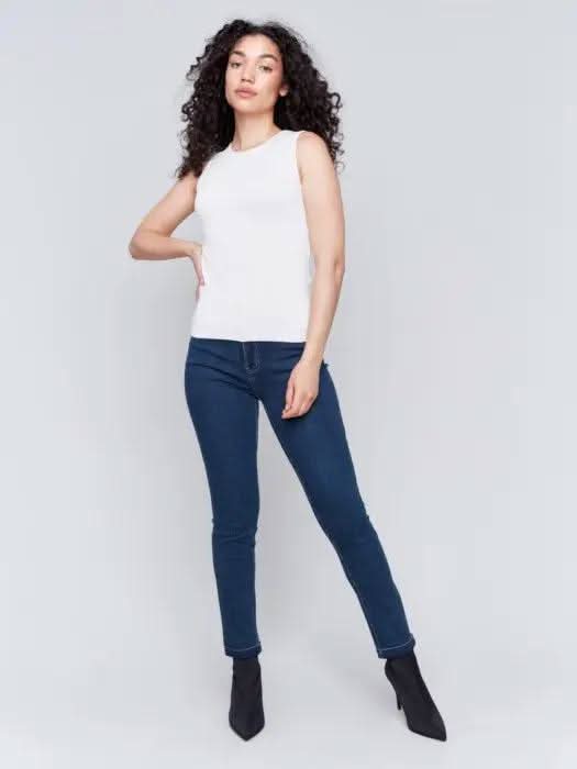 A person with long curly hair, wearing a Charlie B Super Stretch Sleeveless Top, blue jeans, and black ankle boots, stands posing against a plain background.