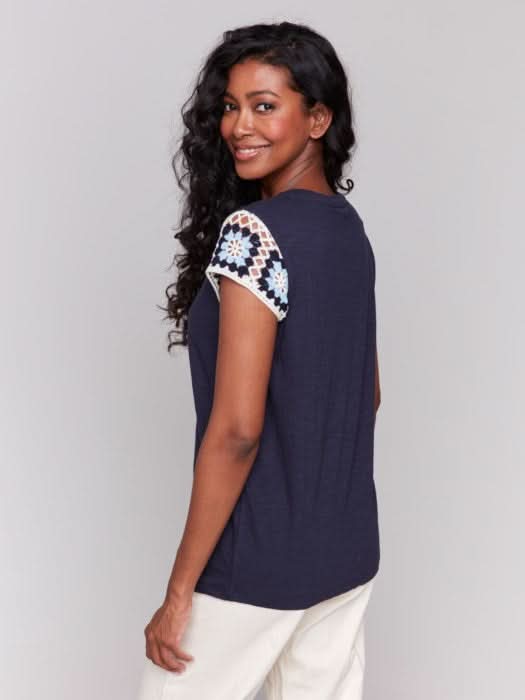 A person with long, wavy hair wears a navy Crochet Cap Sleeve T-Shirt by Charlie B, featuring floral-patterned short sleeves, paired with light-colored pants against a gray background.