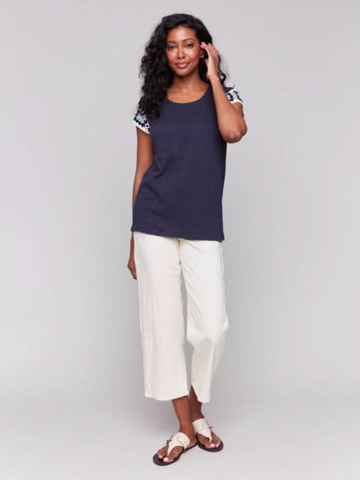A person with long, wavy hair wears a navy Crochet Cap Sleeve T-Shirt by Charlie B, featuring floral-patterned short sleeves, paired with light-colored pants against a gray background.
