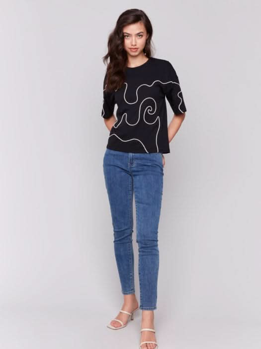 A woman with long hair wears a Charlie B Relaxed Fit Short Sleeve T-Shirt in black, showcasing white abstract line art, paired with blue jeans, ideal for a casual occasion, against a plain gray background.
