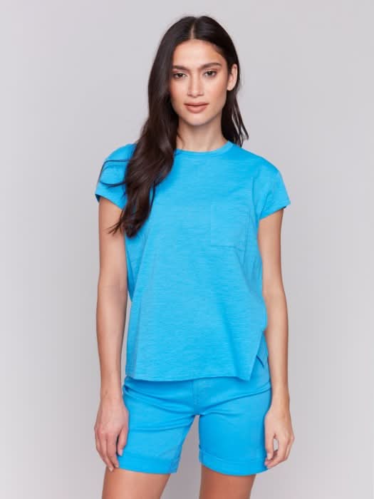 A woman with long dark hair wears the Charlie B Cap Sleeve Tee with Chest Pocket Detail and matching blue shorts made from lightweight fabric, standing against a plain light background.