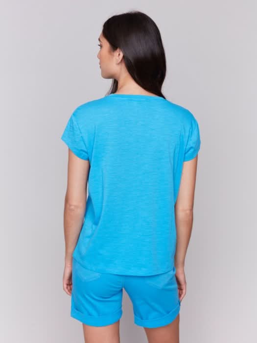 A woman with long dark hair wears the Charlie B Cap Sleeve Tee with Chest Pocket Detail and matching blue shorts made from lightweight fabric, standing against a plain light background.