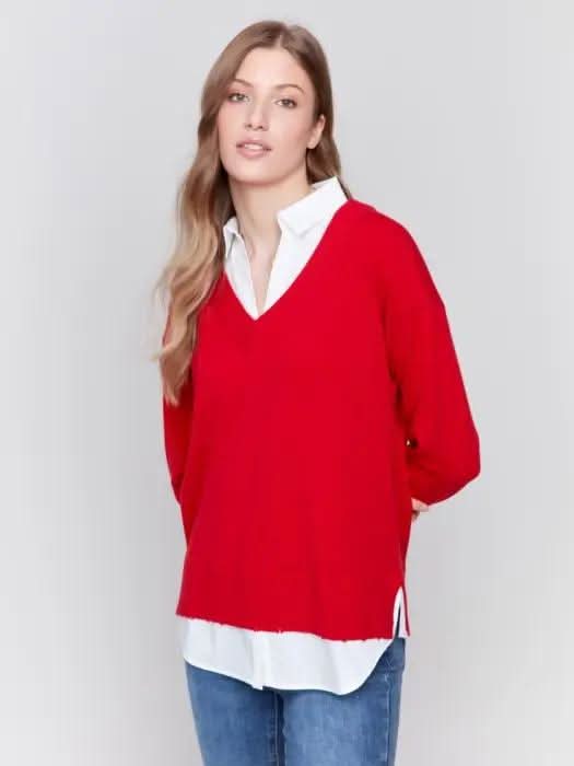 A person with long hair is standing against a gray background, dressed in a red Charlie B V Neck Fooler Sweater with Shirt Collar, complemented by a white shirt, blue jeans, and black boots. This versatile piece introduces a modern flair and effortlessly enhances any wardrobe.
