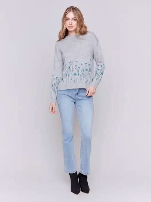 A woman with long hair is wearing the Charlie B Plush Knit Embroidered Flower Sweater featuring gray fabric adorned with blue and green floral details, paired with light blue jeans, creating a look that exudes cozy warmth.