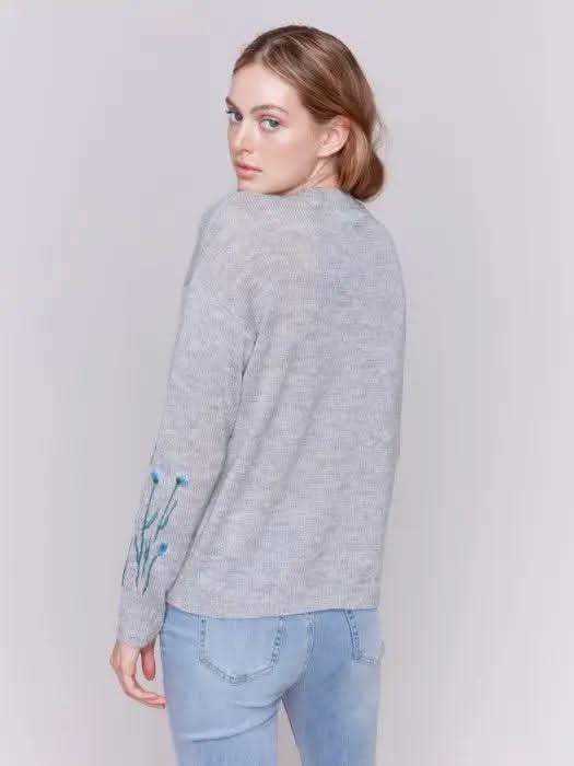 A woman with long hair is wearing the Charlie B Plush Knit Embroidered Flower Sweater featuring gray fabric adorned with blue and green floral details, paired with light blue jeans, creating a look that exudes cozy warmth.