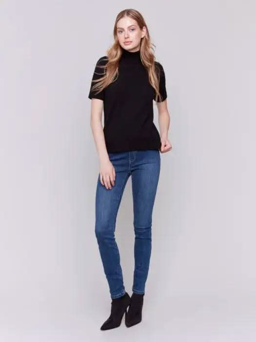 A stylish individual with long hair stands against a plain gray background, donning the Short Sleeve Mock Neck Sweater by Charlie B in black and a pair of comfortably fitting blue jeans.