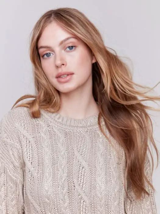 A person with long hair wears a Charlie B Metallic Cable Knit Sweater and white pants, posing against a plain background.