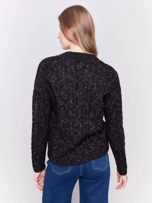 A person with long hair wearing the black Sparkled Yarn Cable Sweater from Charlie B, crafted from ultra-soft material, paired with blue jeans stands against a plain background, looking at the camera.