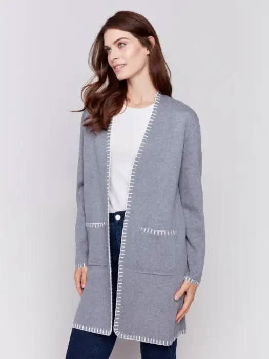 A stylish woman, adorned in a cozy Charlie B Blanket Stitch Cardigan featuring white trim, layered over a white shirt and paired with dark jeans, stands against a plain background.