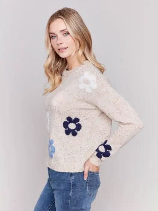 A person with long blonde hair wearing a Charlie B Neps Yarn Knit Sweater adorned with colorful flower patterns and blue jeans stands with hands in pockets against a plain background, perfect for cooler weather.
