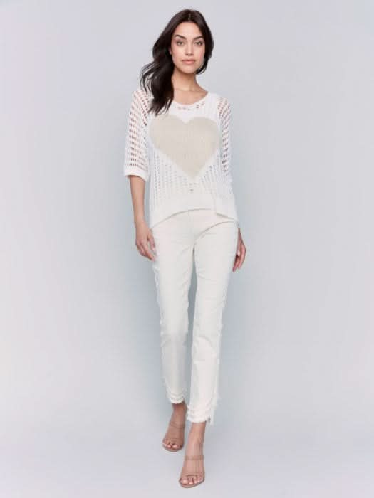 A woman poses against a plain backdrop, wearing the Fishnet V-Neck Sweater with Gold Heart by Charlie B, stylishly paired with light pants.