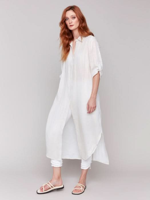 Wearing a Foil Print Linen Blend Tunic Dress by Charlie B over a black top and denim shorts, the woman in a summer outfit poses against a gray background, stylishly finished with white sandals.