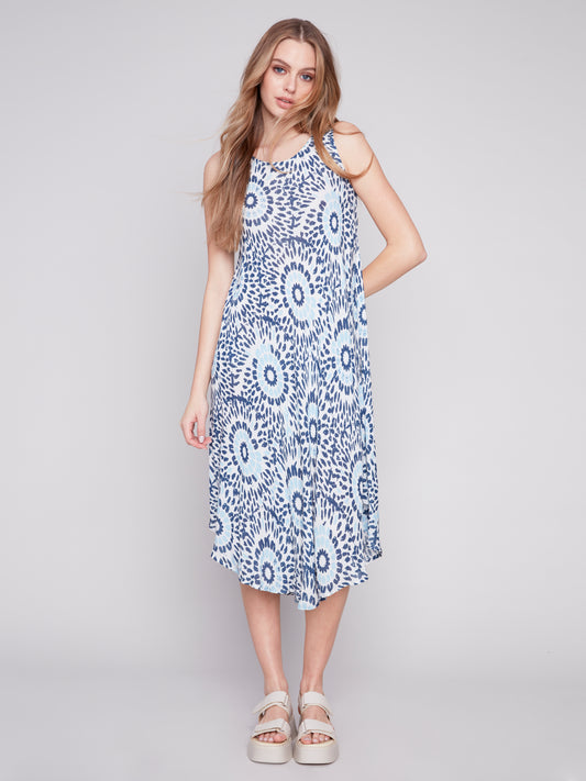 A woman wearing a Charlie B Sahara Print Rayon sleeveless dress in white and blue pattern and white sandals standing against a grey backdrop.