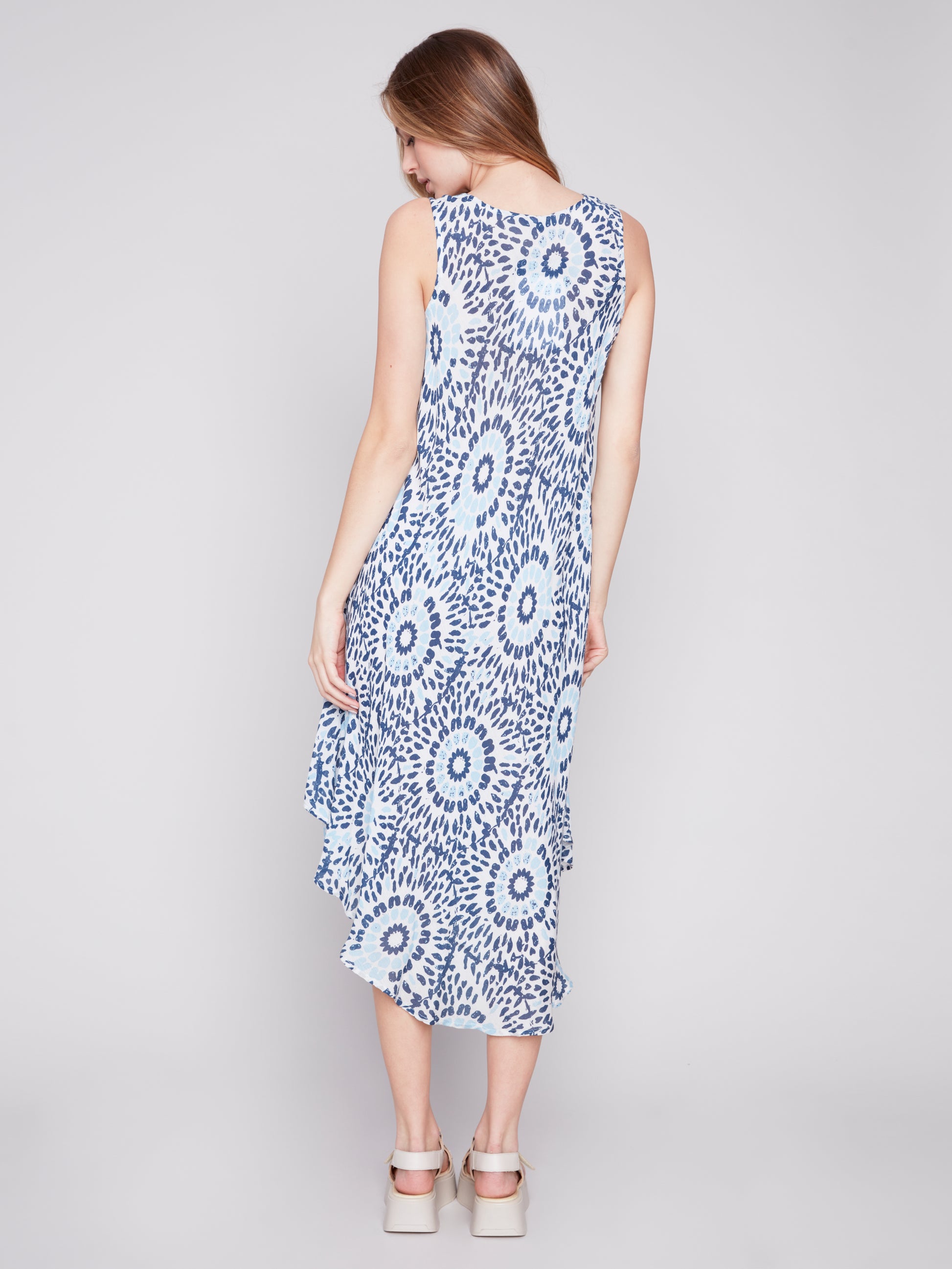A woman wearing a Charlie B Sahara Print Rayon sleeveless dress in white and blue pattern and white sandals standing against a grey backdrop.