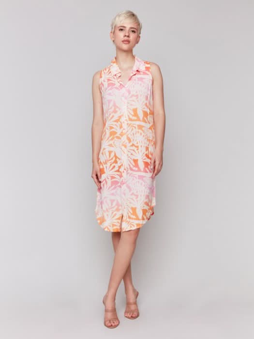 A person with short blonde hair wears the Charlie B Printed Sleeveless Button-Front Dress with a pink and orange leaf pattern and beige heels, standing against a plain light background.