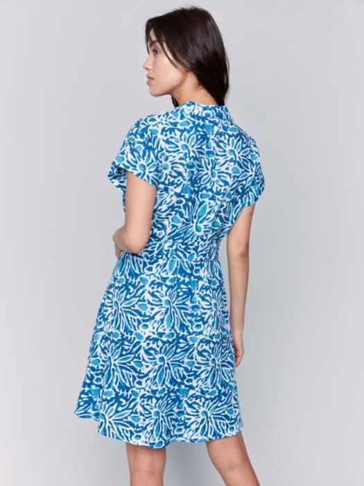 A woman stands against a plain background wearing the Charlie B Printed Short Sleeve Buttoned Dress With Belt in blue and white, styled with white sandals and her hands casually in the dress pockets.