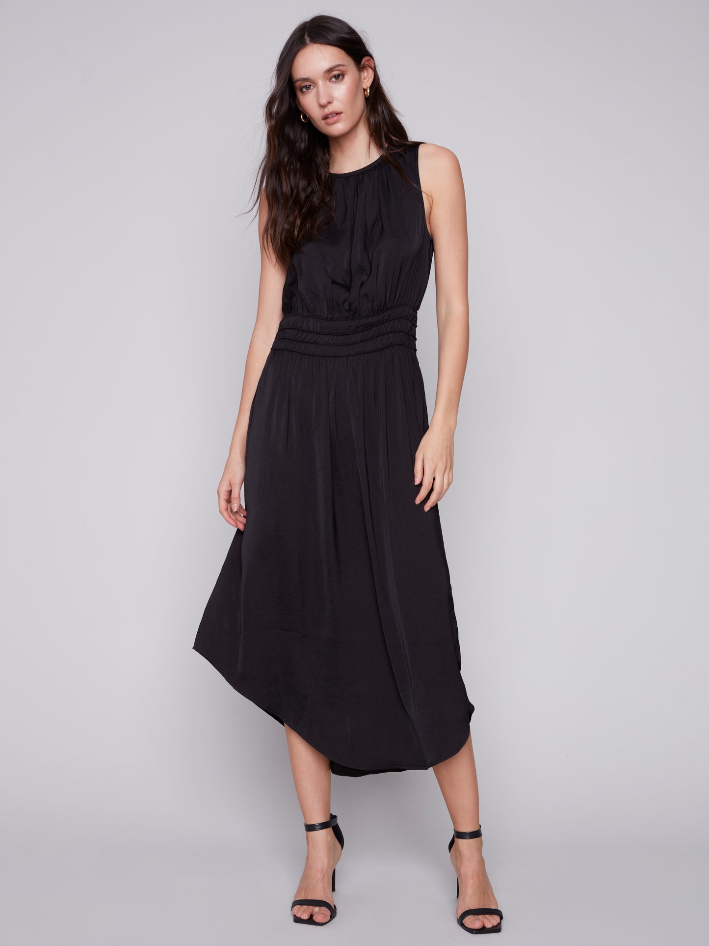A woman in a Charlie B sleeveless black dress with an elastic smocking waist and strappy heels stands against a plain background.