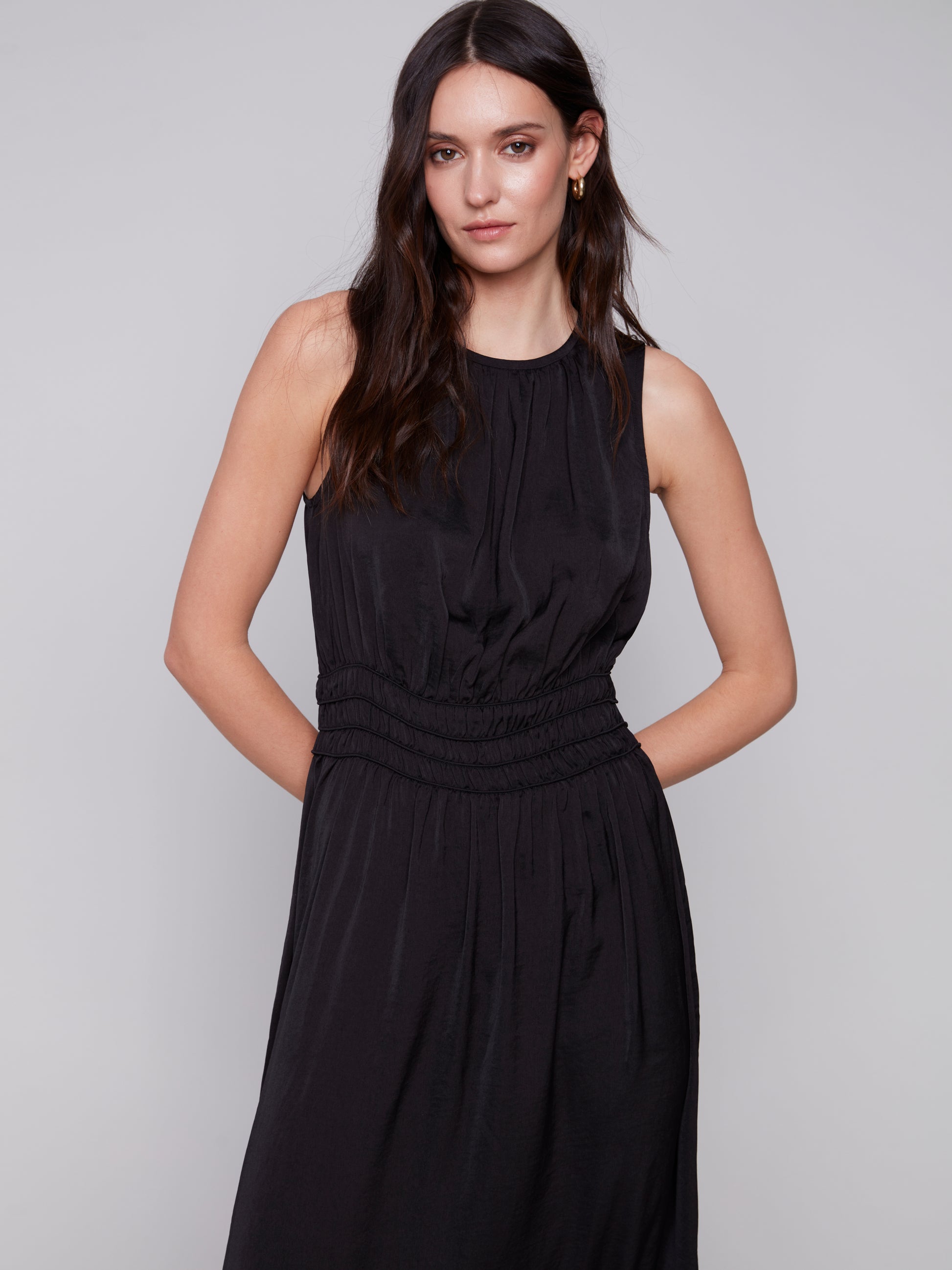 A woman in a Charlie B sleeveless black dress with an elastic smocking waist and strappy heels stands against a plain background.