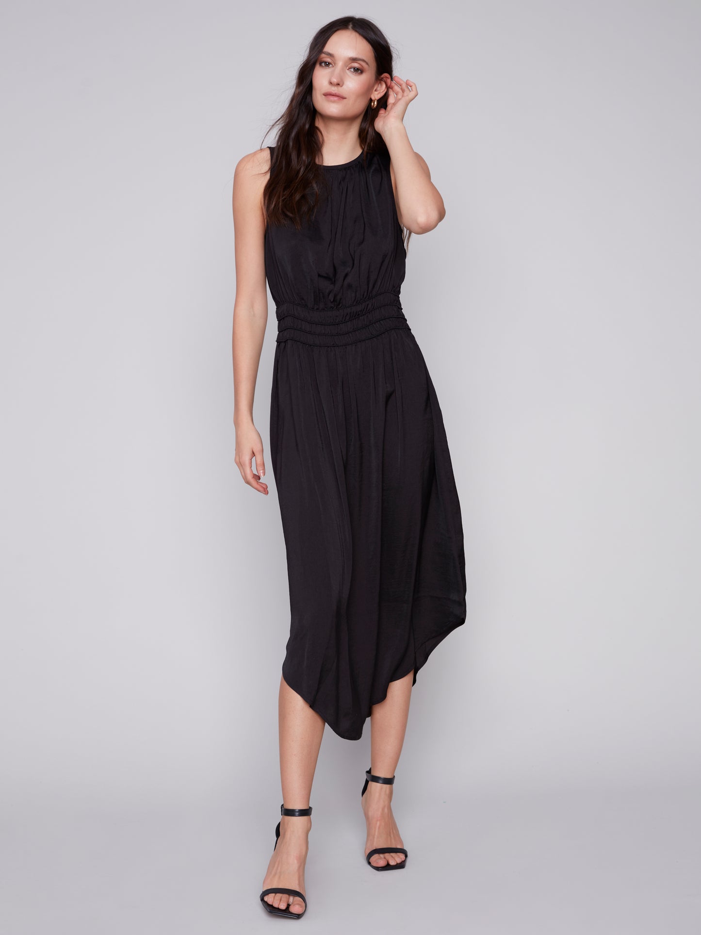 A woman in a Charlie B sleeveless black dress with an elastic smocking waist and strappy heels stands against a plain background.