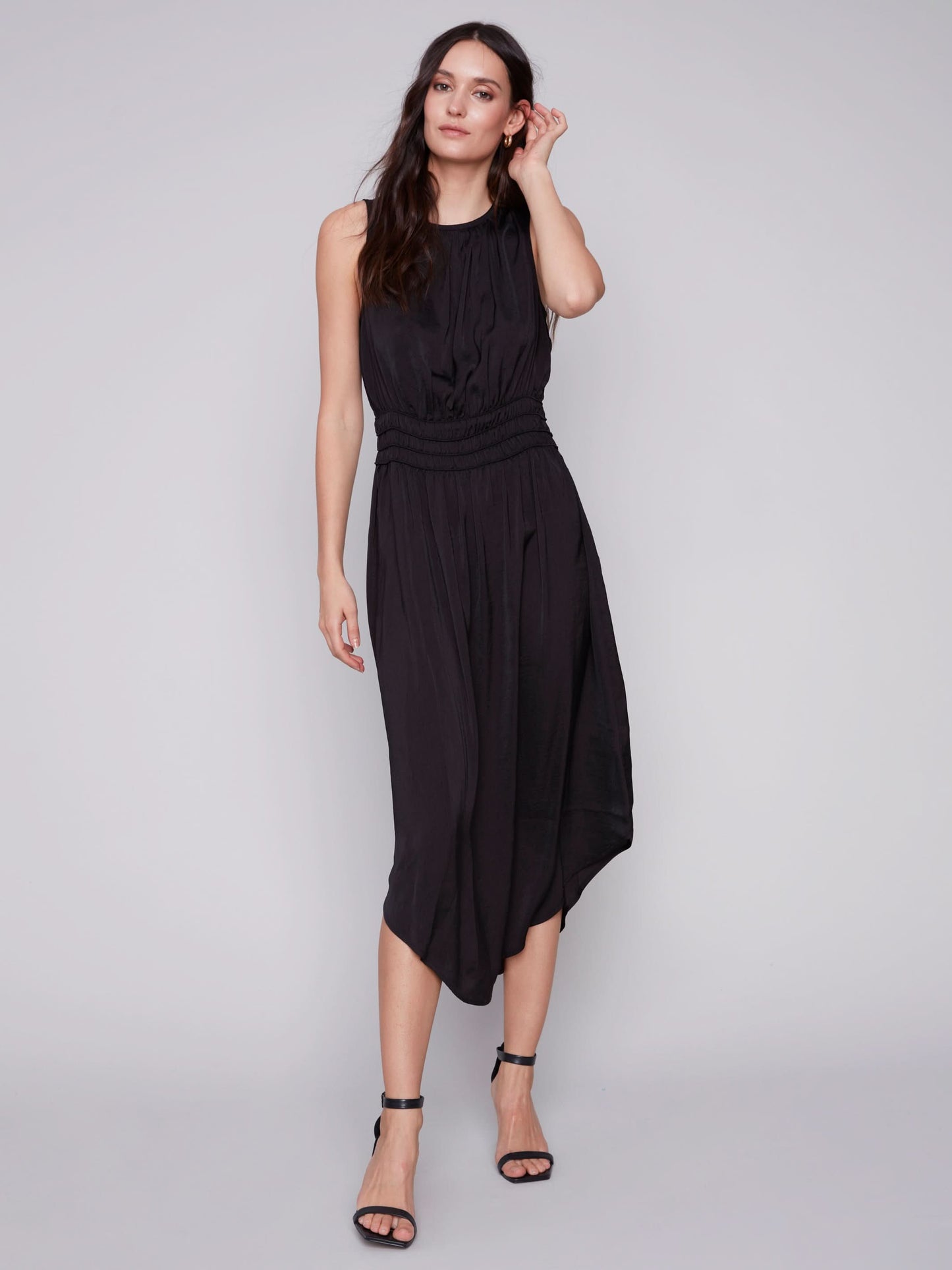 A woman in a Charlie B sleeveless black dress with an elastic smocking waist and strappy heels stands against a plain background.