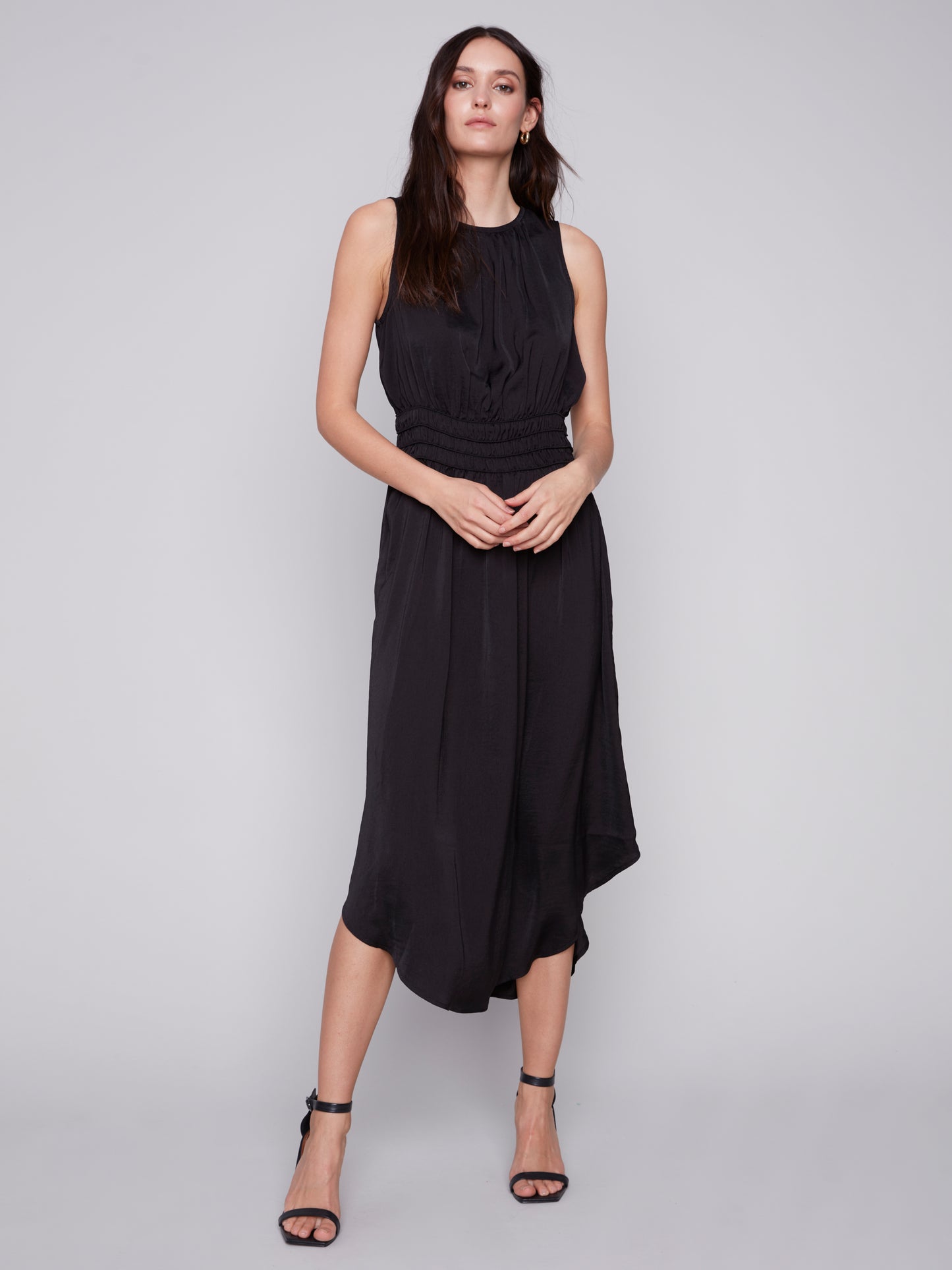 A woman in a Charlie B sleeveless black dress with an elastic smocking waist and strappy heels stands against a plain background.