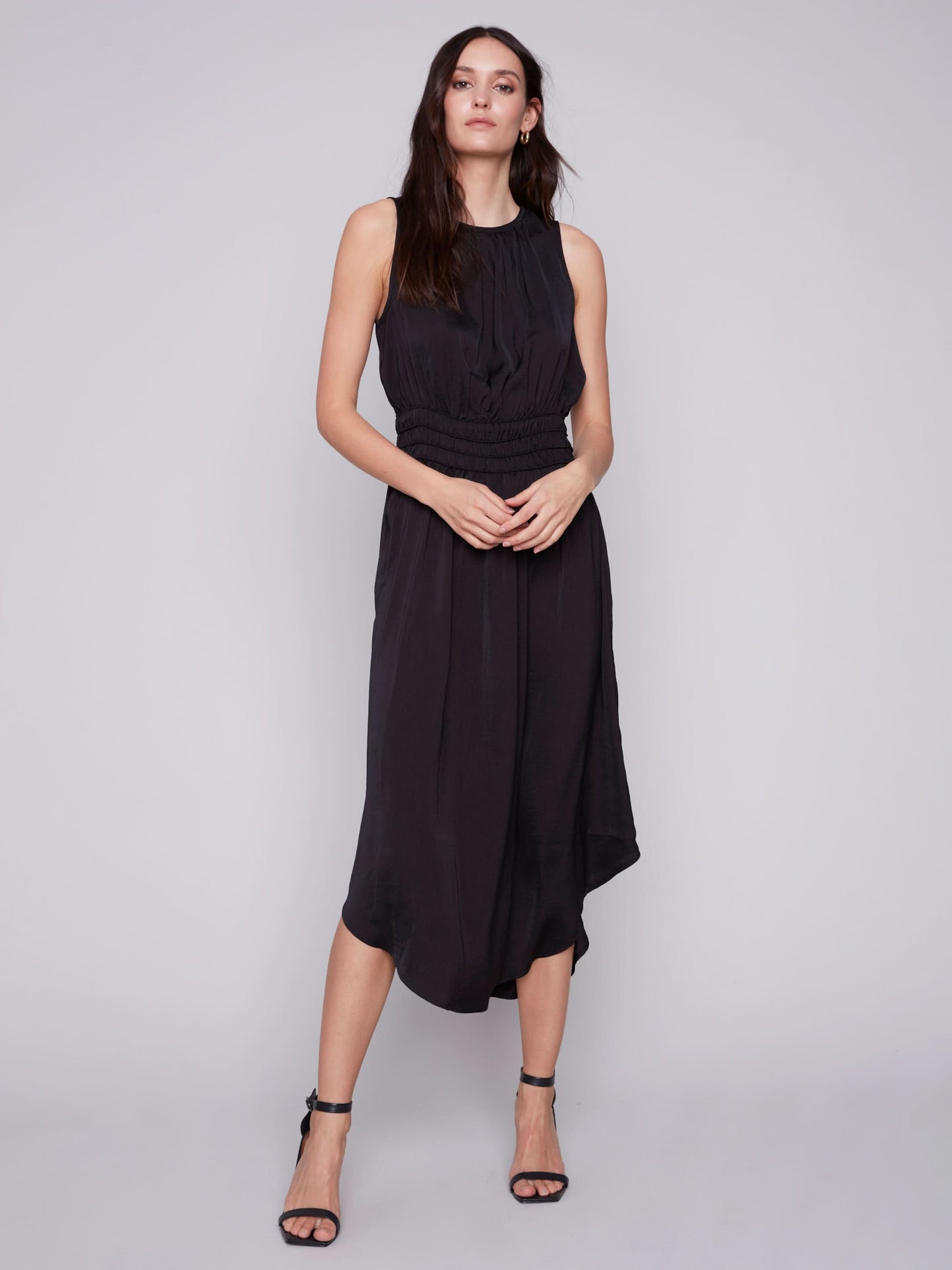 A woman in a Charlie B sleeveless black dress with an elastic smocking waist and strappy heels stands against a plain background.