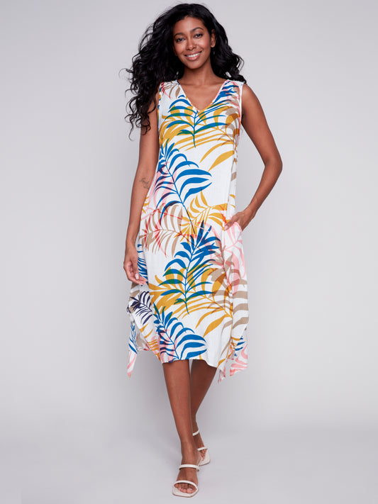 Woman standing in a studio, wearing a colorful tropical print Charlie B midi dress with slits and pockets and beige high heels, smiling at the camera.