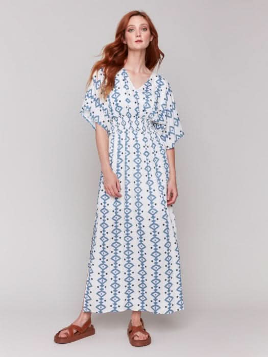 A fashionista in a Charlie B Printed Dolman Sleeve Maxi Dress with Side Slits stands sideways, eyes on the camera, hand on hip, paired with chic white heeled sandals.