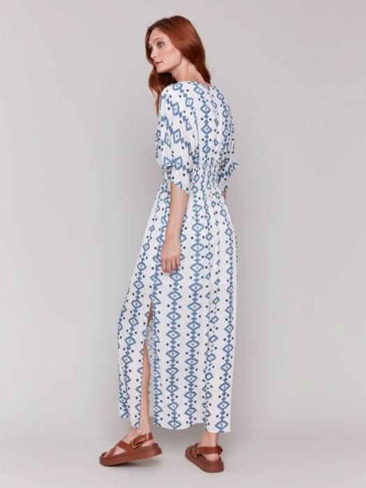 A fashionista in a Charlie B Printed Dolman Sleeve Maxi Dress with Side Slits stands sideways, eyes on the camera, hand on hip, paired with chic white heeled sandals.
